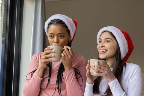 Diverse friends wearing santa hats sipping warm drinks by window for cozy holiday moment