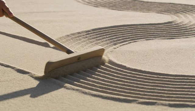 Hand raking zen garden sand with wooden rake creating curved grooves calm minimalist patterns
