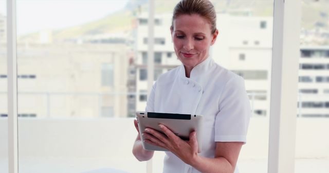 Healthcare professional in white uniform using tablet in a modern office with large windows. Useful for depicting medical technology, healthcare settings, professional roles, and modern medical environments. Ideal for websites, blogs, and presentations related to healthcare, medical technology, and professional workplaces.