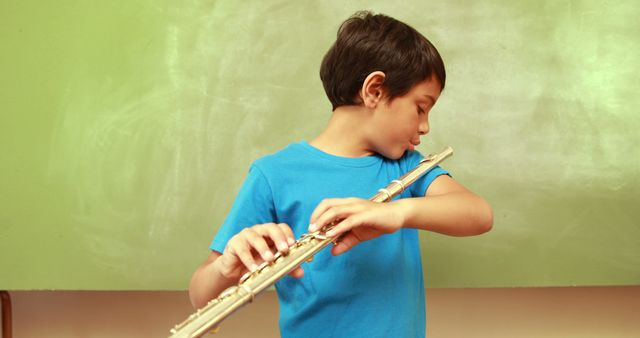 Young boy in blue shirt practicing playing flute indoors with green background. Perfect for educational materials, music-related content, children's activities promotion, or articles on early music education and development.