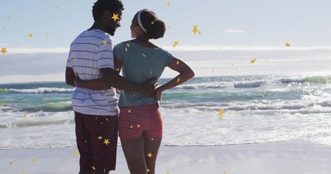 Couple embracing on beach watching ocean waves with gold star confetti falling