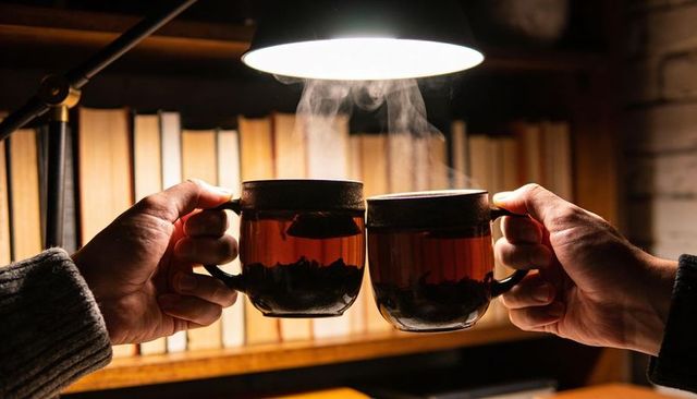 Clinking glass mugs of steaming tea in cozy book-filled study under warm lamp light