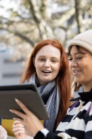Two women using tablet outdoors, smiling and collaborating while wearing beanie and scarf