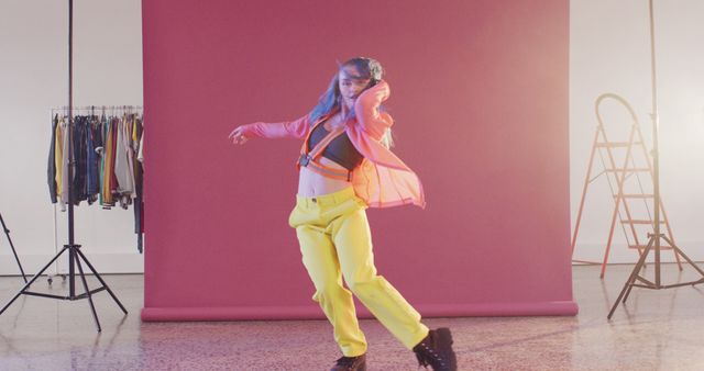 Woman dancing energetically in a brightly colored studio with a pink backdrop. She wears yellow pants and a sheer pink top, adding to the vibrant and trendy mood. Ideal for use in articles or advertising related to dance, fashion, youth culture, or creative industries. Fits well for promotions on social media, lifestyle blogs, and fashion magazines.