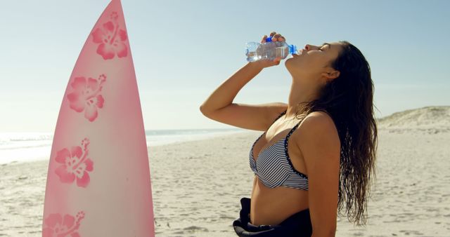 Young biracial woman hydrates after surfing at the beach. She stands by her pink surfboard, enjoying the sunny seaside ambiance.