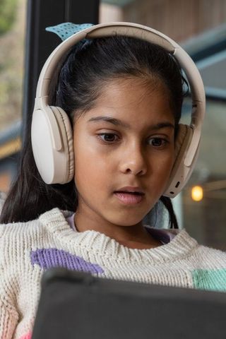 Indian girl wearing beige wireless headphones using tablet for remote learning by window