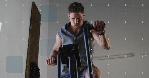 Focused man pedaling air bike in garage gym wearing sleeveless hoodie and fitness tracker