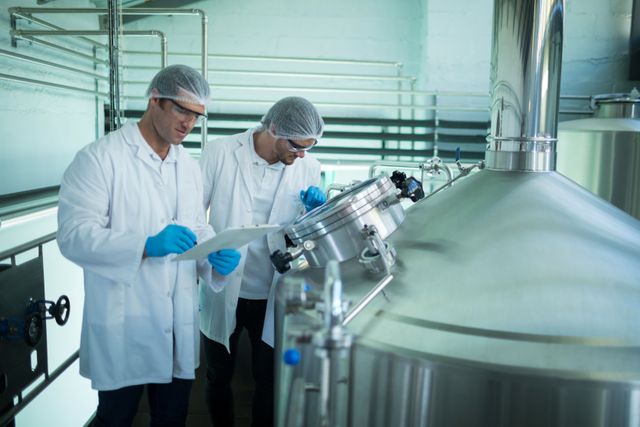Two scientists in lab coats, gloves, and hairnets are discussing near a large storage tank in an industrial factory. This image can be used for themes related to scientific research, industrial processes, quality control, manufacturing, and technology advancements.