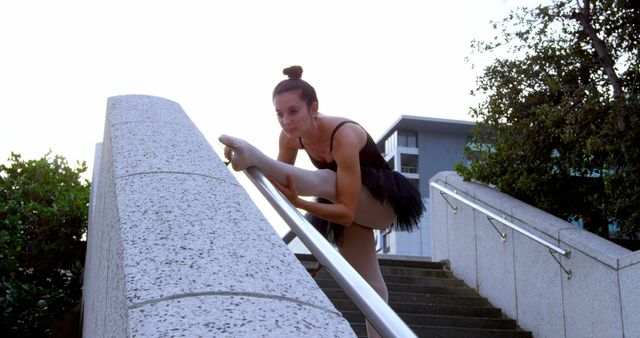 Female ballet dancer stretching on a staircase railing in an urban environment. She wears a black tutu and demonstrates flexibility and strength. Perfect for use in content related to dance, fitness, urban practices, or artistic expressions.