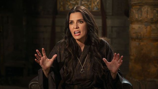 Medieval storyteller in dark robe gesturing with ringed hands and pendant, ancient stone interior