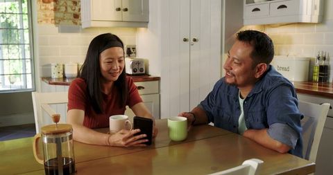 Asian couple sharing coffee and smartphone at sunlit rustic kitchen table, morning chat