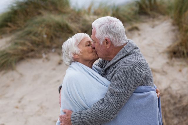 Elderly couple sharing a tender kiss on a sandy beach, wrapped in blankets to stay warm. The intimacy and affection showcased make this image perfect for websites, brochures, and campaigns promoting senior living, romance, and outdoor activities for older adults. Ideal for illustrating themes of long-term relationships, love, and companionship in marketing materials.