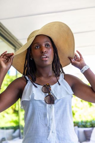 African woman adjusting wide-brim straw hat on patio wearing white ruffled top