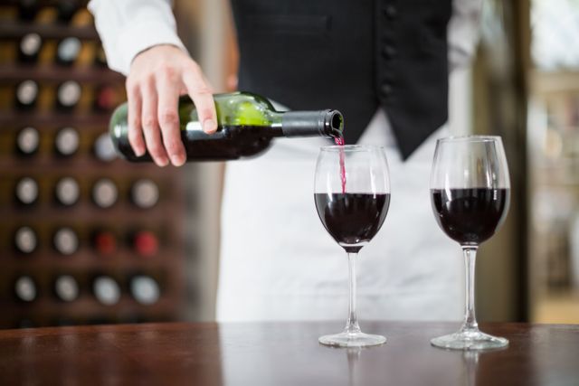 Waiter in uniform pouring red wine into two wine glasses on a wooden table. Ideal for use in content related to restaurants, fine dining, hospitality industry, wine tasting events, and luxury dining experiences.