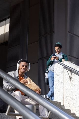 African american college friends sitting on campus steps with headphones and smartphone
