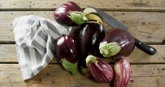 Eggplants of various sizes and shapes arranged on rustic wooden table with kitchen knife and striped cloth next to them. Ideal for use in cooking blogs, farm-to-table commercials, healthy eating promotions, and food-related articles.