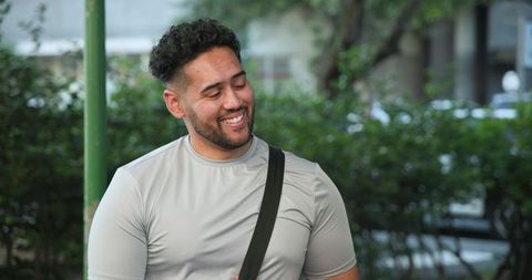 Smiling man standing on urban sidewalk with crossbody strap and green hedges