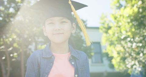 Young girl wearing graduation cap and tassel smiling while standing in sunlit schoolyard