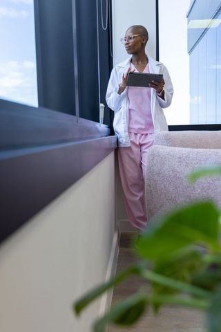 Contemplative african clinician in pink scrubs holding tablet by window