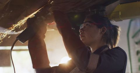 Female mechanic grinding metal under vehicle with safety gear