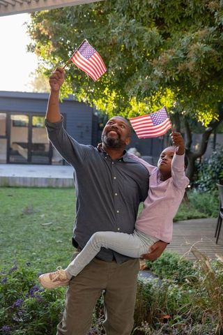 African american father holding daughter waving american flags in backyard celebration