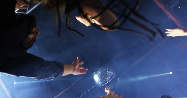 Group of friends reaching towards a shiny disco ball under bright lights in a nightclub setting. Ideal for use in content related to nightlife entertainment, partying, social gatherings, and festive atmospheres.