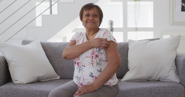 Senior woman sitting on couch indoors, showing her arm after getting vaccinated, indicating health consciousness and preventive care. Ideal for healthcare campaigns, vaccination awareness, and healthy living promotions.