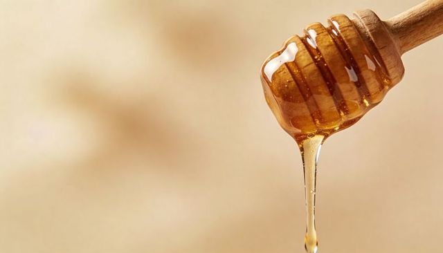 Golden Honey Dripping from Wooden Dipper on Warm Beige Background, Closeup Drop