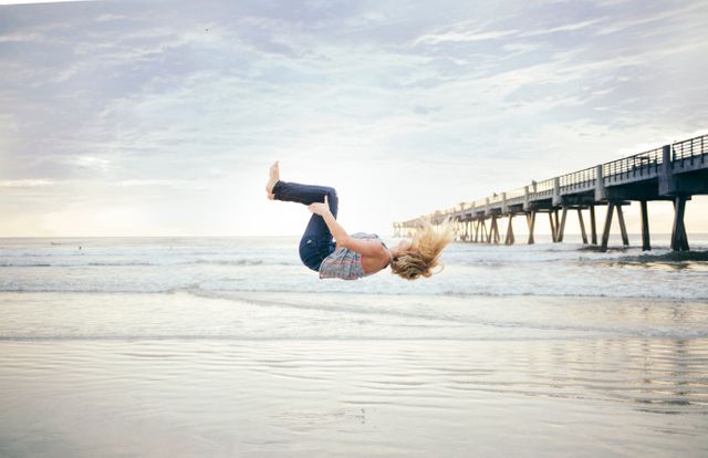 Woman performing backflip by the ocean near a pier during sunrise. Suitable for themes related to fitness, outdoor activities, beach outings, and athletic endeavors. Can be used for motivational posters, fitness advertisements, travel blogs, and social media content promoting a healthy lifestyle by the sea.
