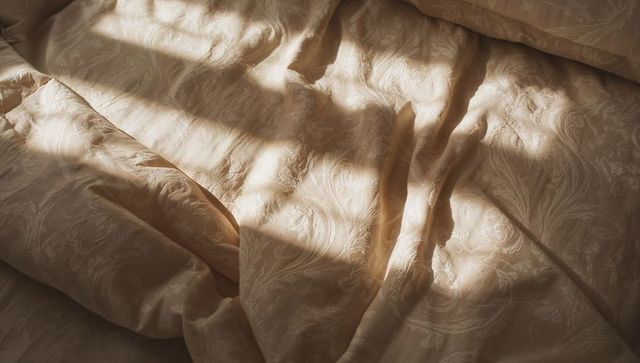 Beige damask duvet unfolding in warm window light with soft pillow and shadows