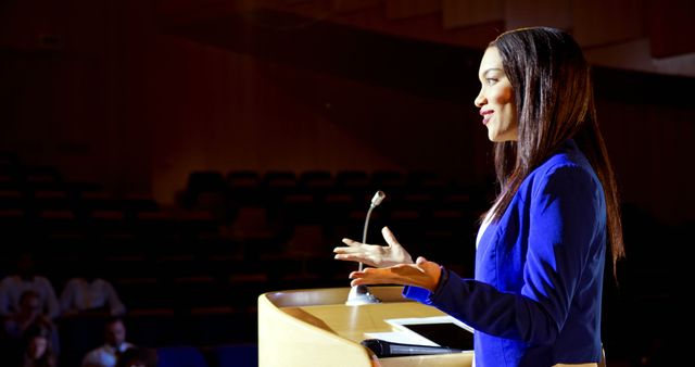 Young woman dressed in blue blazer is giving a confident presentation at the podium with a microphone and tablet in an auditorium. Useful for illustrating themes related to public speaking, education, business conferences, leadership development, and professional presentations.
