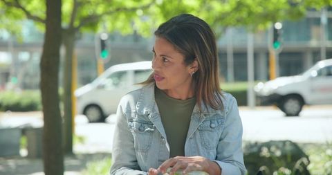 Woman wearing denim jacket sitting on bench in leafy urban plaza looking sideways in sunlight