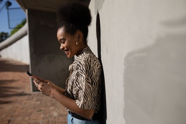 Side view of a happy biracial woman enjoying free time in a city on a sunny day, using a smartphone, smiling leaning against a wall, wearing blue jeans and zebra print shirt.