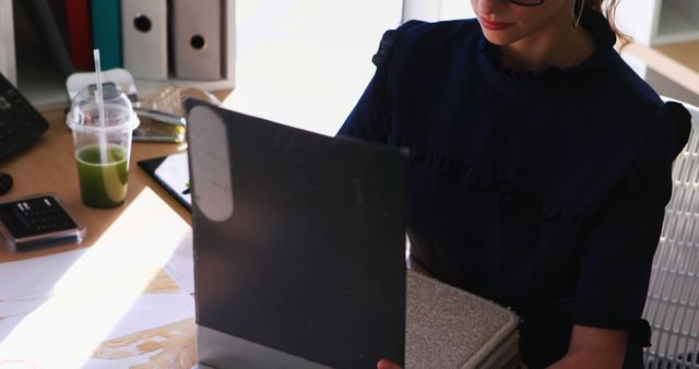 Woman working at office desk with various material texture samples and a green smoothie. Suitable for concepts related to design, creativity, architecture, professional work environment, and business tasks.