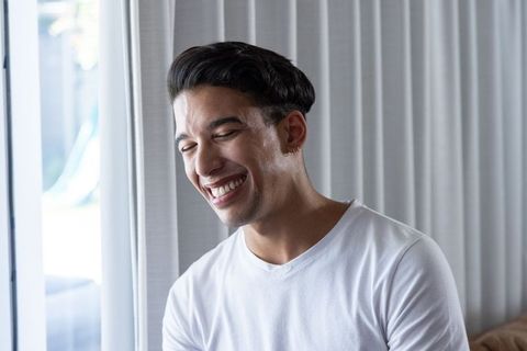 Young african american man laughing by window wearing white t-shirt, casual home lifestyle portrait