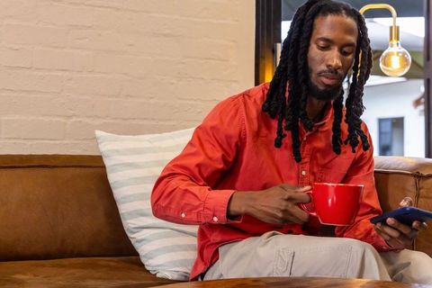African american man wearing red shirt sipping coffee and scrolling smartphone on leather sofa