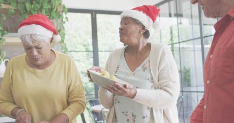Senior women wearing Santa hats sharing plate of apples in sunlit family holiday kitchen