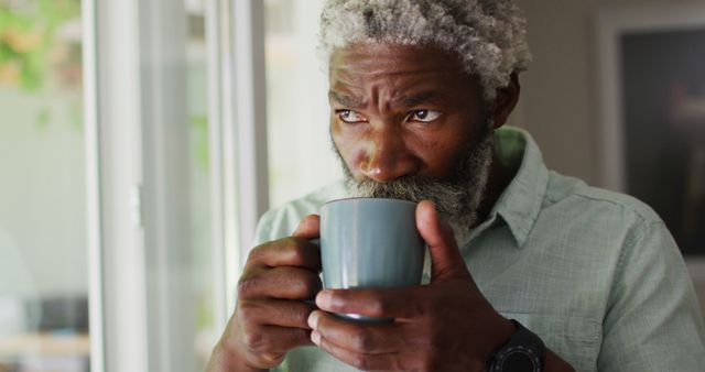 Portrait of african american senior man with beard drinking coffee looking out of window and smiling. staying at home in isolation during quarantine lockdown.