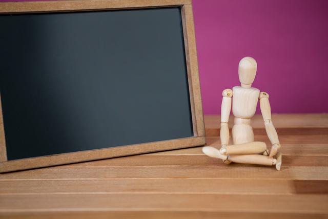This image features a wooden mannequin sitting on a wooden floor next to a blank chalkboard against a purple background. Ideal for illustrating concepts of education, learning, teaching, creativity, art, and presentations. Suitable for use in educational content, blogs, websites, and marketing materials focused on creativity and classroom environments.