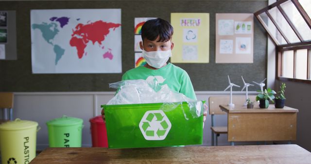 Young boy holding a green recycling bin full of plastic waste in a classroom, promoting eco-friendly and sustainable practices. Posters on the wall and recycling bins in the background suggest an educational setting focused on environmental lessons. Ideal for campaigns or materials on environmental awareness, school sustainability programs, or Earth Day initiatives.