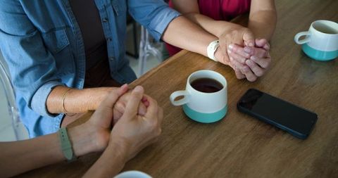 Diverse female friends holding hands over coffee, senior companionship and support