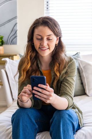 Young woman sitting on sofa using smartphone, smiling in cozy daylight living room
