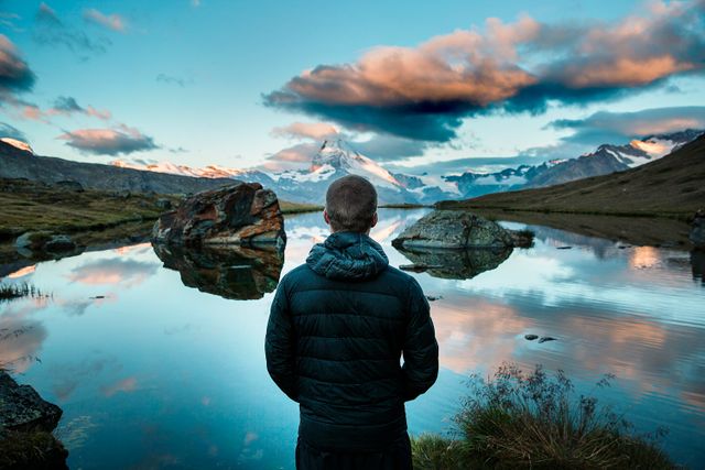 Man standing at edge of calm lake, gazing at breathtaking mountain scenery with dramatic clouds overhead. Ideal for travel blogs, outdoor adventure articles, nature conservation campaigns, and inspirational posters. Conveys peace, exploration, and the beauty of nature.