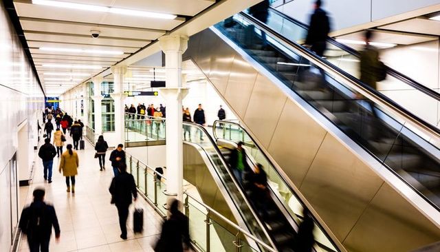 Busy urban transit concourse with moving commuters, escalators, glass and steel interior