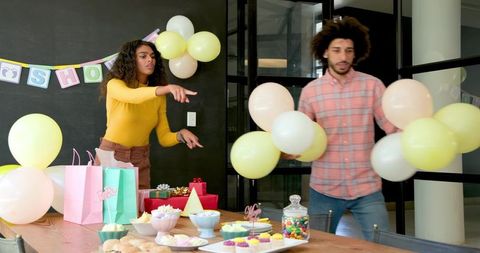 Diverse friends arranging pastel balloons and banner for baby shower with cupcake table