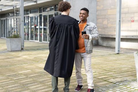 Smiling friends congratulating graduate in gown on campus courtyard using smartphone