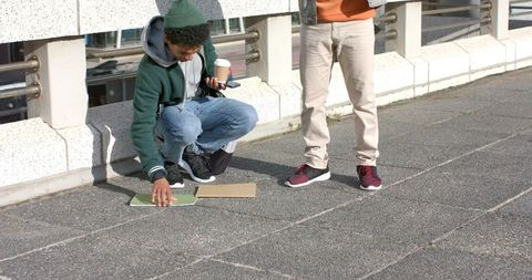 Young men picking up green spiral notebook on rooftop walkway holding coffee and phone