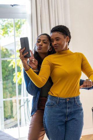 Black woman and south asian friend taking playful selfie in sunlit living room, casual fashion