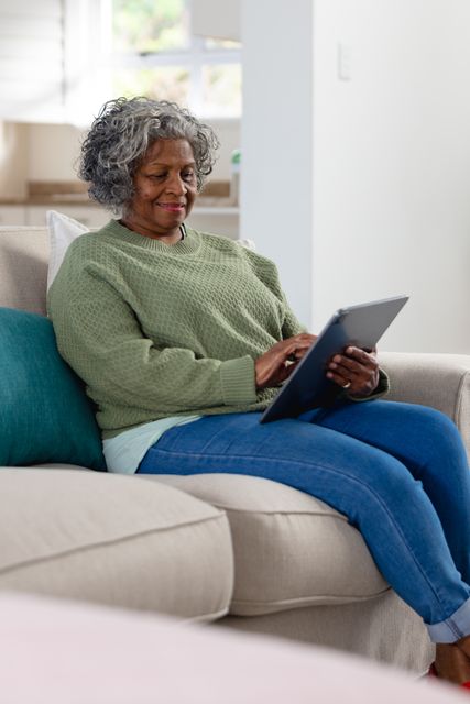 Vertical of happy senior african american woman using tablet sitting in sofa living room, copy space. Retirement, communication, technology, inclusivity and senior lifestyle concept.