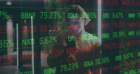 Man checking stock tickers on glass wall in modern lobby using smartphone